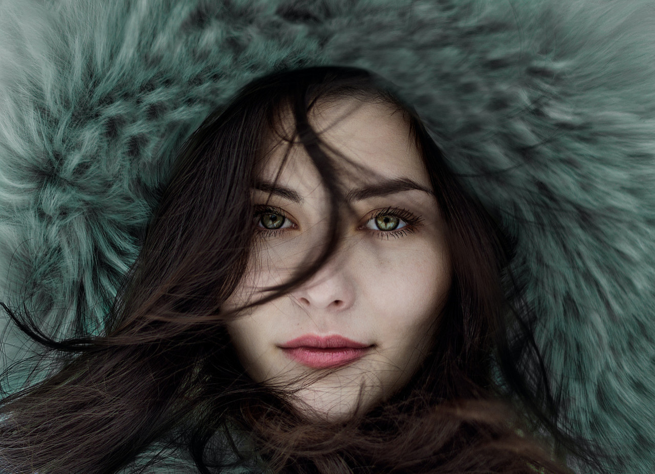 *belle peau hiver*  une femme aux yeux vert et avec une capuche fourrée se tient dans le froid de l'hiver.