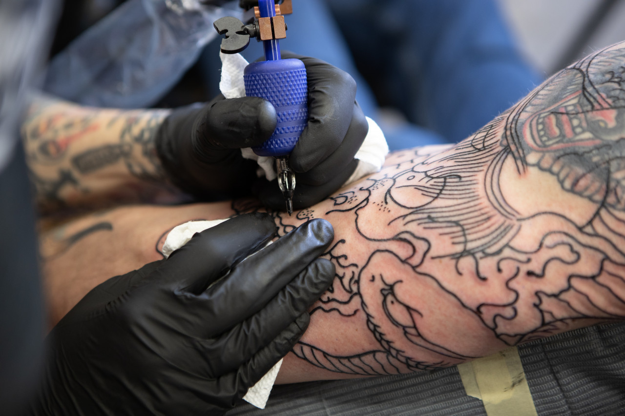 *danger tatouage*  Un tatoueur s'affaire sur un bras, avec des gans noirs aux mains.