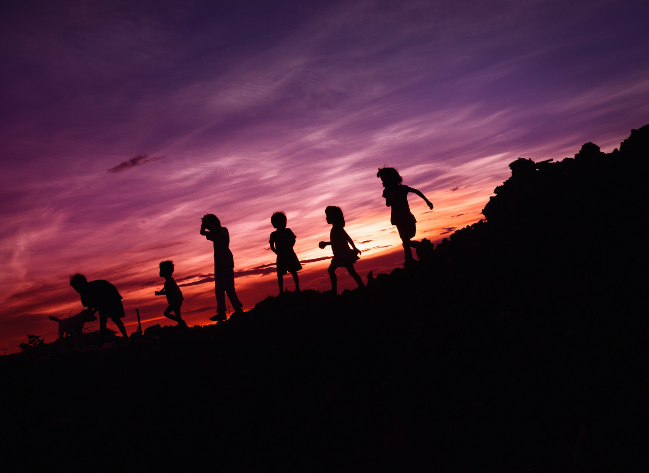 *balade au coucher de soleil*  Les ombres d'un groupe d'enfants et parents marchent sur un chemin montagneux avec un ciel orange et violet en fond.