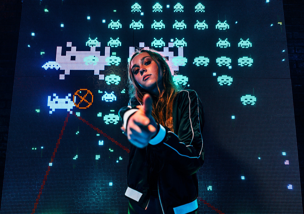 *femme gameuse*  Jeune femme qui se tient devant un écran de jeu vidéo où se joue space invaders.