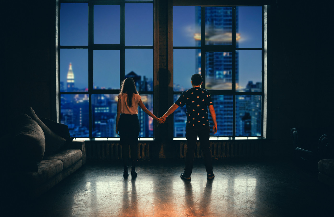 *couple*  Un couple dans un appartement se tient la main, ils regarde la ville éclairée depuis la fenêtre. La nuit est tombée
